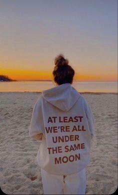 a woman standing on top of a sandy beach next to the ocean wearing a white hoodie