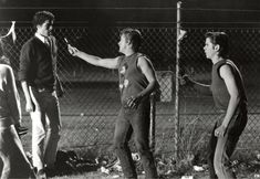 three young men standing next to each other in front of a chain link fence, talking