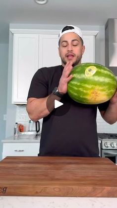 a man holding up a large watermelon in his hand while standing next to a kitchen counter