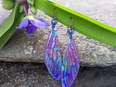 a pair of blue and pink earrings sitting on top of a rock next to a purple flower