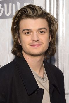 a young man with long hair wearing a black jacket and tan shirt is smiling at the camera