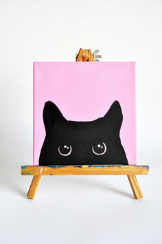 a black cat on a pink background is sitting on a wooden easel and it's eyes are drawn