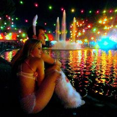 two people sitting on the edge of a body of water with lights in the background