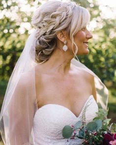 a woman in a wedding dress holding a bouquet and looking off to the side with trees behind her