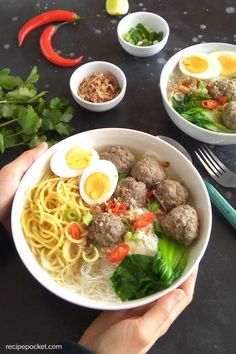 someone holding a bowl of noodles with meatballs and veggies in it on a table