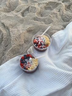 two bowls of fruit are sitting on a towel in the sand, with strawberries and blueberries