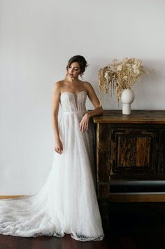 a woman standing in front of a wooden table wearing a white wedding dress and holding her hand on the side