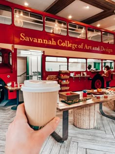 a hand holding a coffee cup in front of a red double decker bus at savannah college of art and design