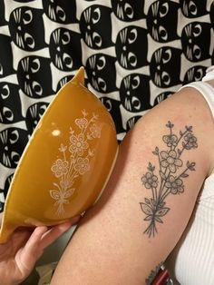 a woman with a tattoo on her arm holding a yellow bowl and flower arrangement in it