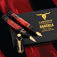 two fountain pens sitting on top of a black box next to red and gold foil