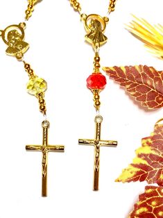 two gold cross pendants with red and yellow beads on a white surface next to autumn leaves