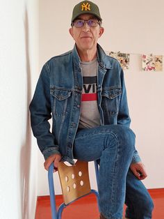 an older man sitting on top of a chair wearing a baseball cap and jeans with his legs crossed