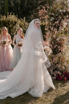 three women in dresses and veils are walking through the grass with flowers behind them