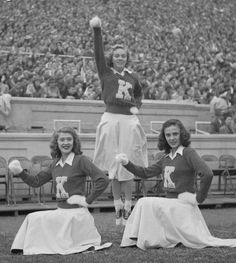 three women in cheerleader uniforms standing on the sidelines at a football game, with one holding her arms up