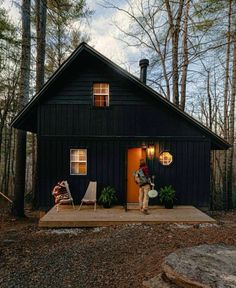 a small black cabin in the woods with two lawn chairs and one person standing outside