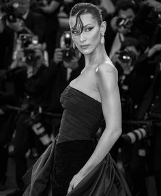 a woman in a black dress posing for the camera with cameras around her and people behind her