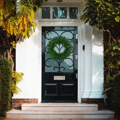 a black door with a green wreath on it's glass front door, surrounded by trees and bushes