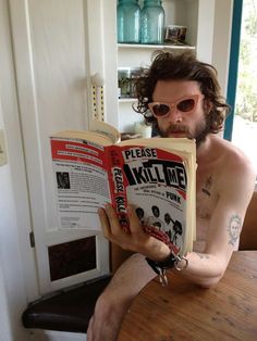 a shirtless man sitting at a table holding an open book in front of his face