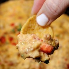 a hand holding a tortilla chip with cheese and vegetables on it in front of a bowl of dip