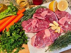 raw meats and vegetables on a cutting board next to lemons, carrots, parsley