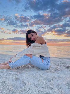 a beautiful woman sitting on top of a sandy beach next to the ocean at sunset