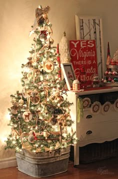 a small christmas tree in a bucket next to a white dresser and red sign that says yes virginia there is a little bit of cheer on it