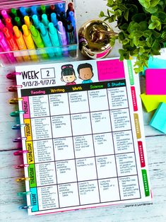 a weekly planner with markers and pens on a white wooden table next to a potted plant