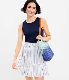 a woman in a blue top and white skirt holding a handbag smiling at the camera