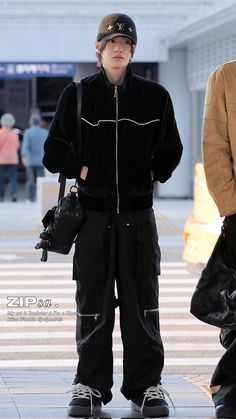 a young boy standing on the sidewalk with his hands in his pockets, wearing black pants and a baseball cap