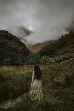 a woman in a white dress walking through a lush green field under a cloudy sky