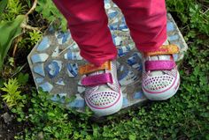 a child's feet wearing pink and yellow shoes standing on a stepping stone in the grass