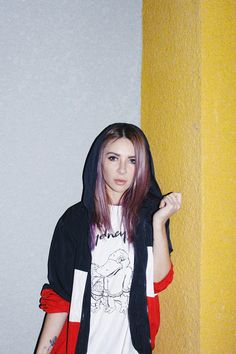 a young woman with pink hair wearing a black and white hoodie standing in front of a yellow wall