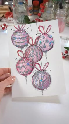 a hand holding up a card with ornaments on it and ribbon around the balls in pink