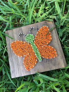 a string art butterfly sitting on top of a piece of wood in the green grass