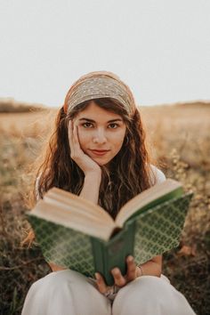 a woman sitting in a field reading a book with her hands on her face and looking at the camera