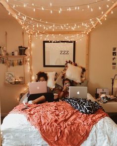 two people sitting on a bed with laptops in front of their faces and lights strung across the room