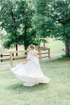 a woman in a white dress is walking through the grass with trees and fence behind her