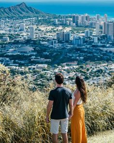 a man and woman standing on top of a grass covered hill looking at the city