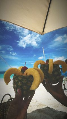 someone holding up two bananas in front of an umbrella on the beach with blue sky and white clouds