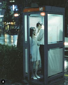 two people standing in a phone booth on the side of a street at night time