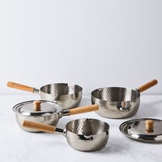 four stainless steel pots and pans with wooden handles on a marble countertop in front of a white wall