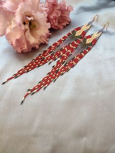 a pair of red and gold beaded earrings next to pink carnations on a blue cloth