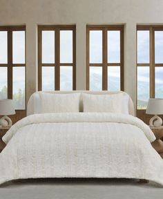 a bed with white comforter and pillows in front of three windows