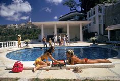 two women sitting on the edge of a swimming pool in front of a large house