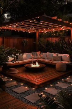 an outdoor fire pit surrounded by wooden decking with lights on the roof and trees in the background