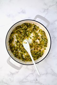 a white spoon in a pot filled with green vegetables and sauce on top of a marble counter