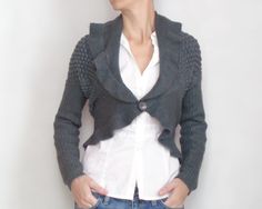 a woman standing in front of a white wall wearing a gray cardigan sweater and jeans