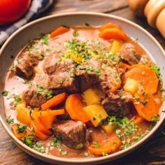 a bowl filled with stew and carrots on top of a wooden table next to vegetables