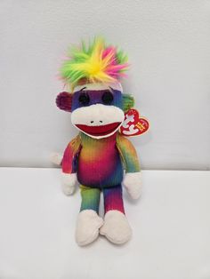 a stuffed monkey with multi colored hair and a tag on its ear sitting against a white wall