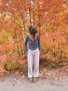 a woman standing in front of trees with leaves on the ground and her hands behind her head